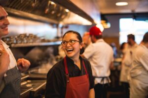 A woman chef laughing in a busy restaurant kitchen